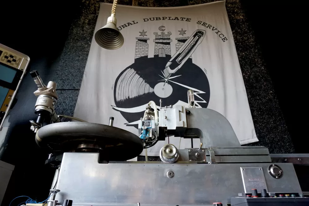 An image of the lacquer cutter from Kommunal Dubplate Service in front of a huge banner in the background with their logo on it.